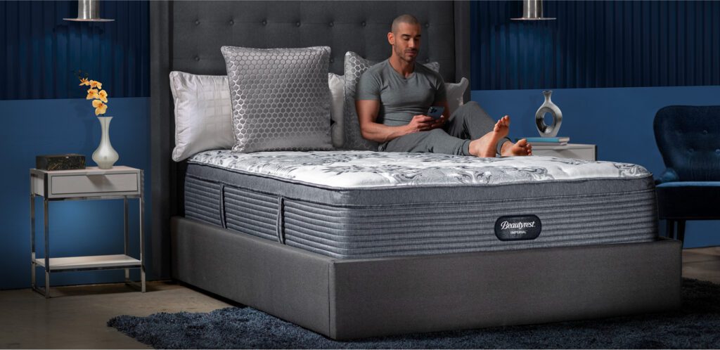 Man looking at his phone while sitting on Beautyrest Imperial Lux mattress in a bedroom.