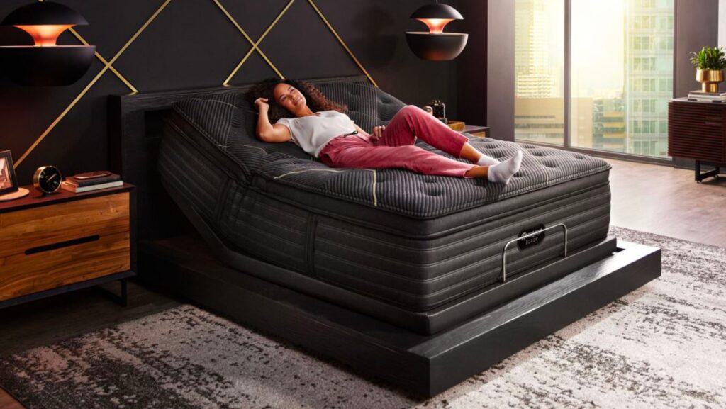 Woman lying on a Beautyrest Black mattress in a well-lit bedroom with a window overlooking a city skyline.