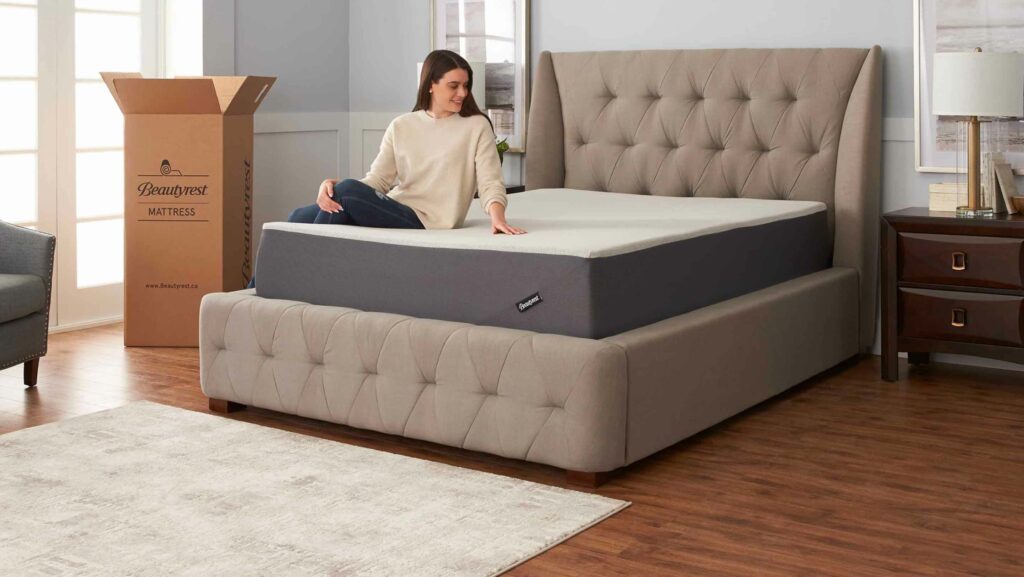 Woman sitting on a Beautyrest Mattress in a Box in a well-lit bedroom with a window and a cardboard box in front.